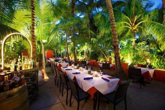 Outdoor restaurant with tables surrounded by tropical palm trees at night.