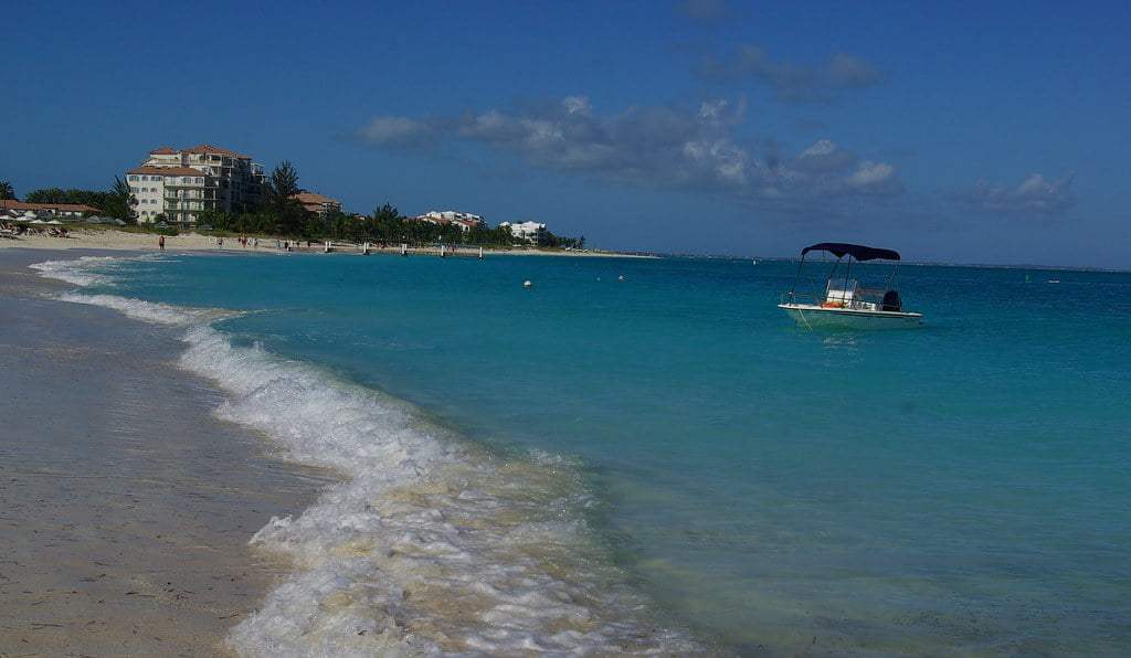 Beach with turquoise water, a boat, and buildings in the distance under a blue sky.