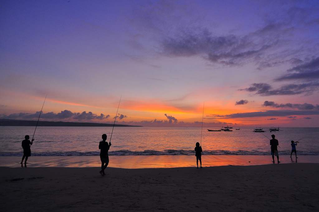 Silhouetted people fishing on a beach at sunset with boats in the distance.