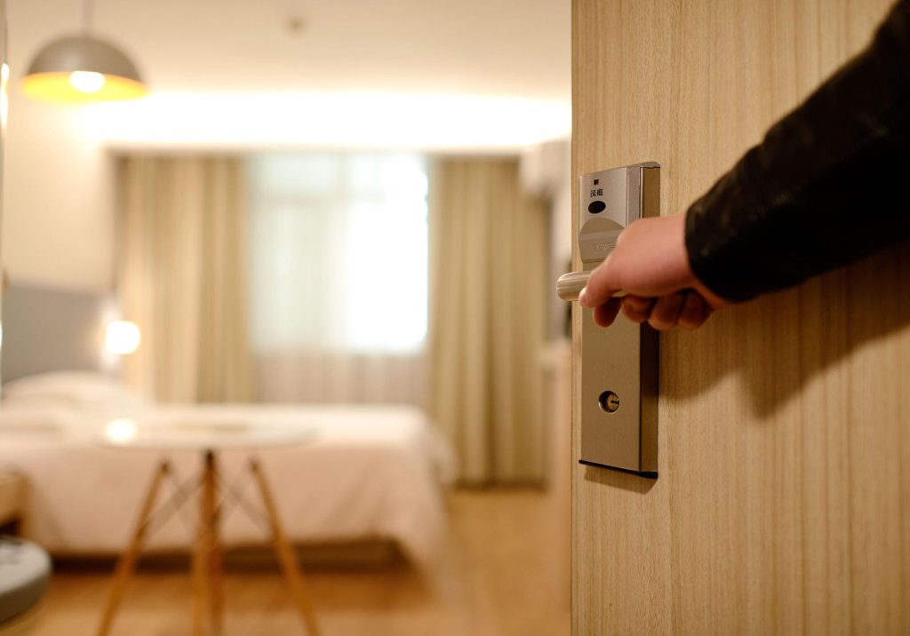 Hand opening door to a bright, modern hotel room with a bed and curtains.