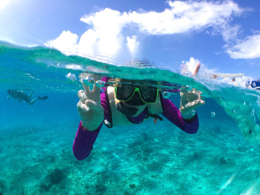 Person snorkeling underwater with peace sign, wearing purple shirt and mask.