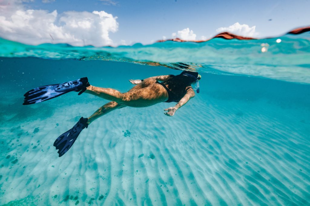 Person snorkeling with flippers in clear blue water near sand.