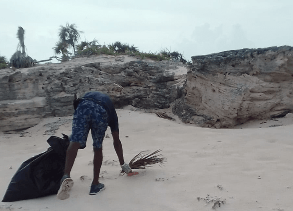 Person cleaning beach with large garbage bag near rocky area.