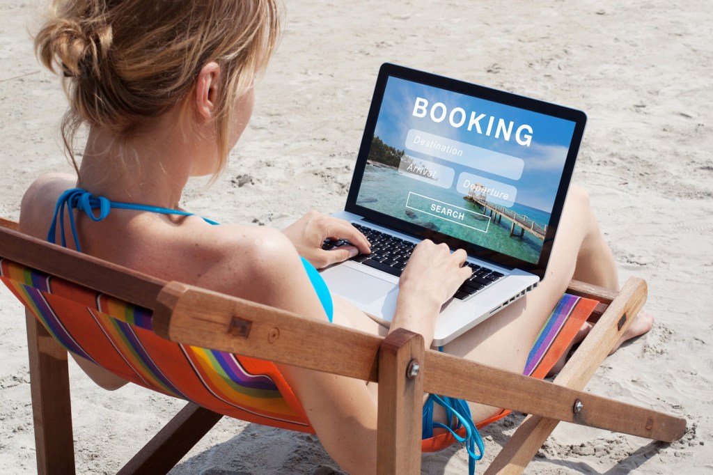 Woman in a beach chair using a laptop with a booking website on the screen.