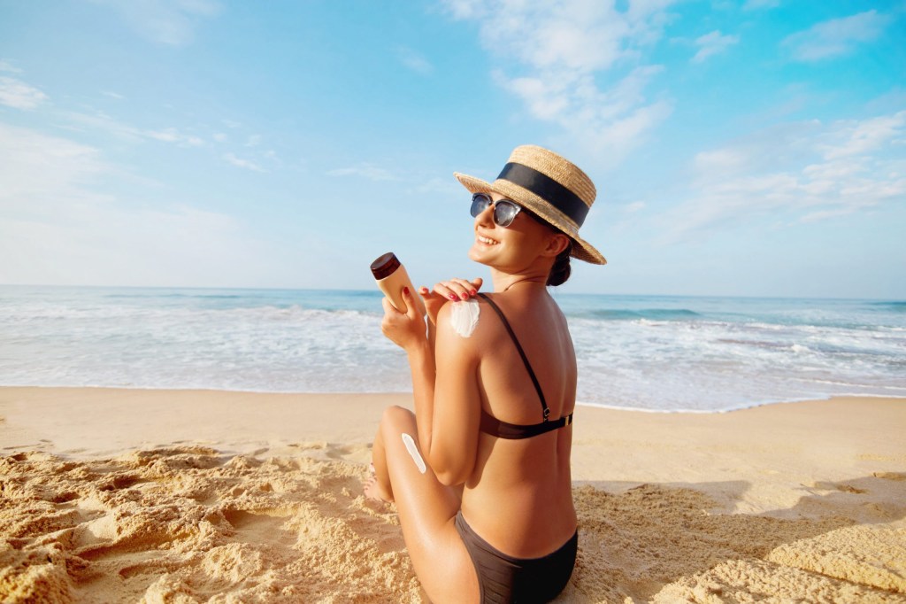 Woman in black bikini and hat applying sunscreen on a sunny beach.