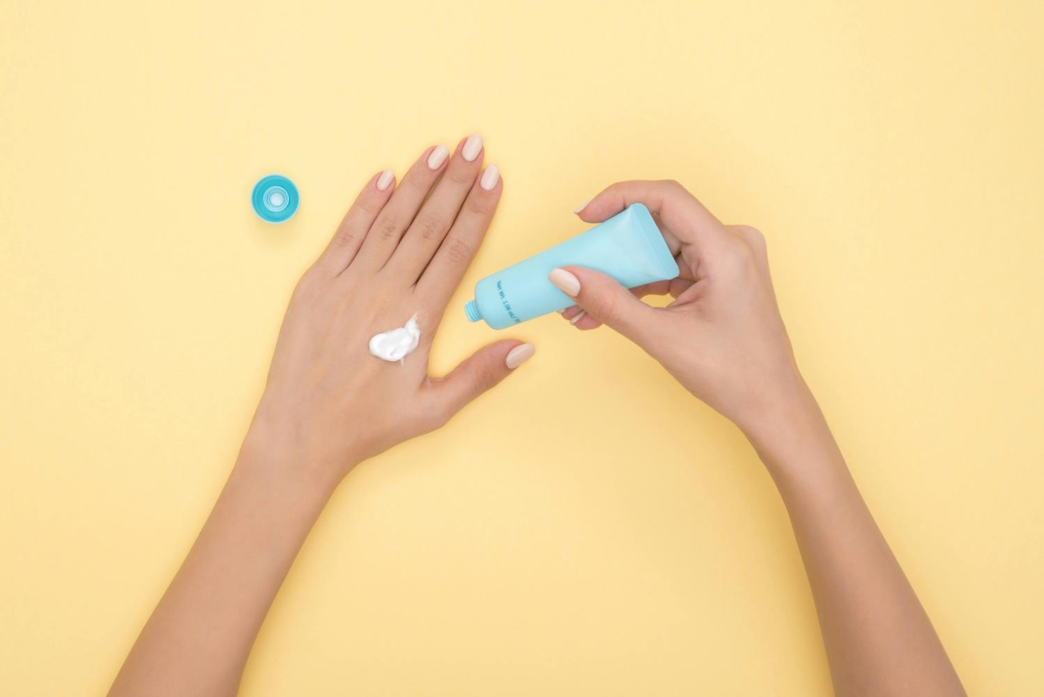 Hands applying cream from a blue tube on a yellow background.