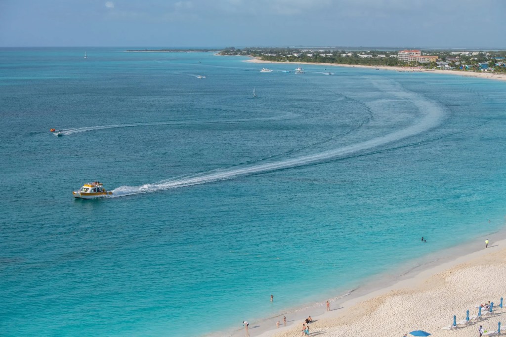 Beach view with a boat creating a wake on the ocean, people on the shore, and a distant coastline.