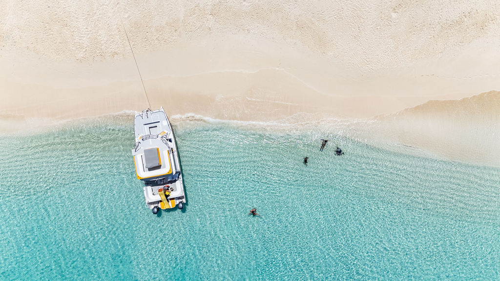 Aerial view of a boat anchored near a sandy beach with people swimming in clear blue water.