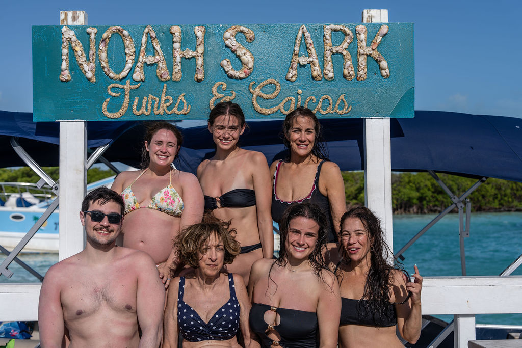 Group of people in swimsuits posing by a sign on a boat labeled 'Noah's Ark Turks & Caicos'.