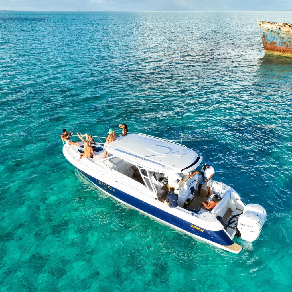 People on a white motorboat in clear blue ocean near a rusted shipwreck.
