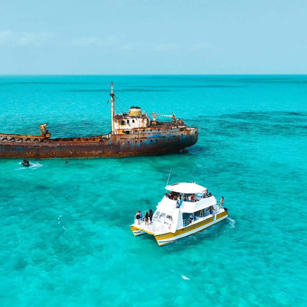 Boat near a rusty shipwreck in clear turquoise water under a blue sky.