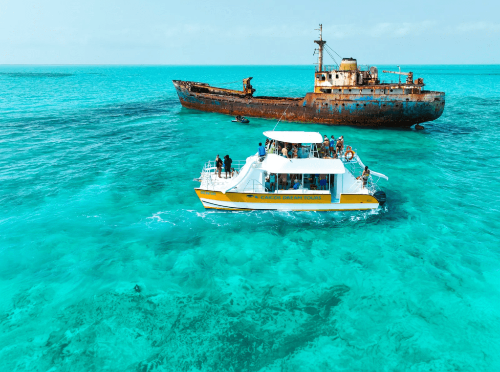People on a boat near a rusty shipwreck in clear blue water.