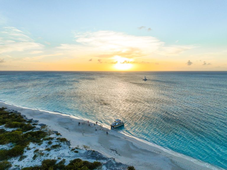 Sunset over ocean with boat on sandy beach, people nearby.