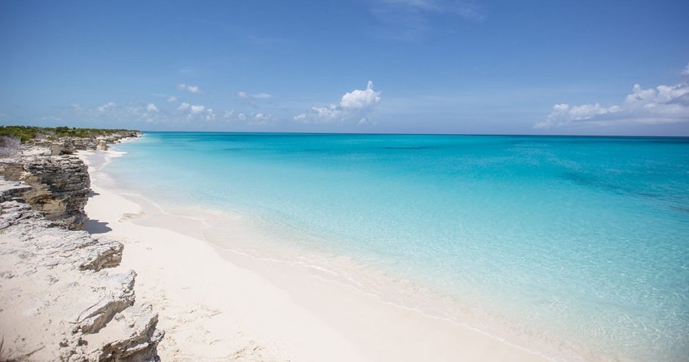 Tropical beach with turquoise water, white sand, and rocky shoreline under a clear blue sky.