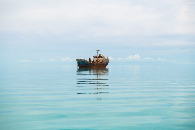 Rusty ship floating on calm sea under cloudy sky.