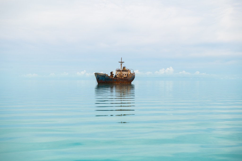 Rusty ship floating on calm sea under cloudy sky.