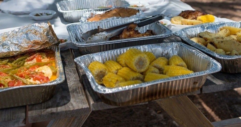 Aluminum trays with grilled food, including corn and vegetables, on a picnic table.