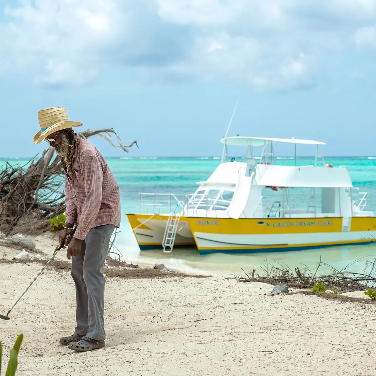 Man with straw hat sweeping sand near a yellow boat by the ocean.