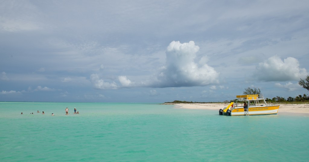 People swimming near a beach with a yellow boat on turquoise water and cloudy sky.