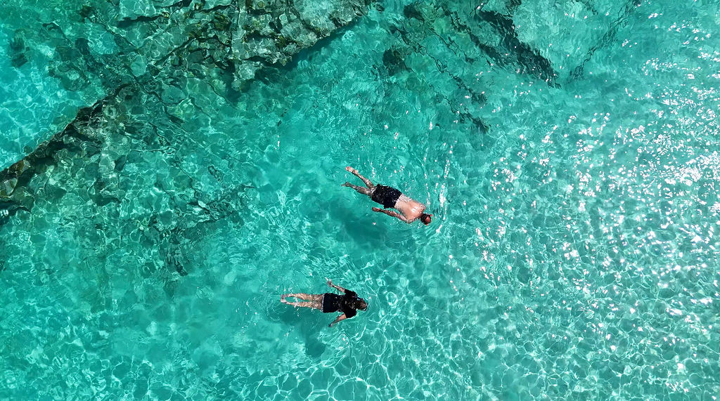 Two people swimming in clear turquoise water with visible rocks below.
