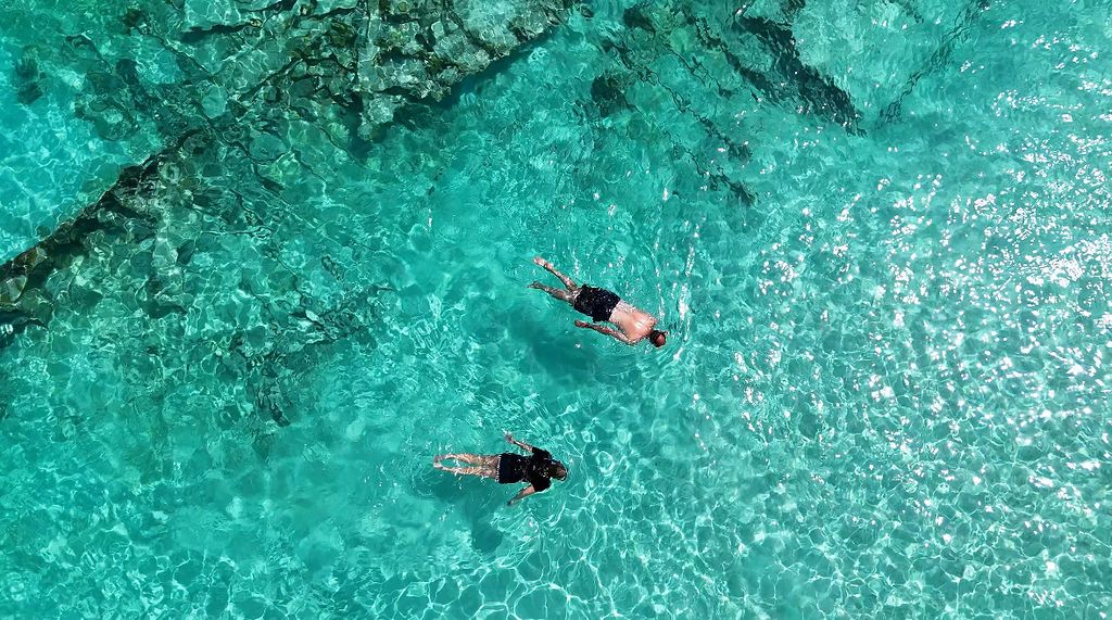 Two people swimming in clear turquoise water with visible rocks below.
