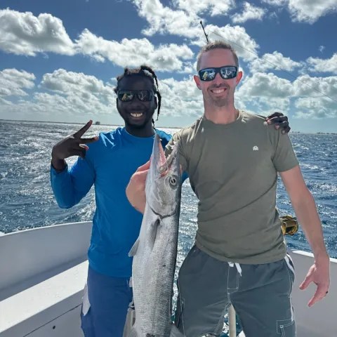 Two men on a boat, one holding a large fish, smiling under a cloudy sky.