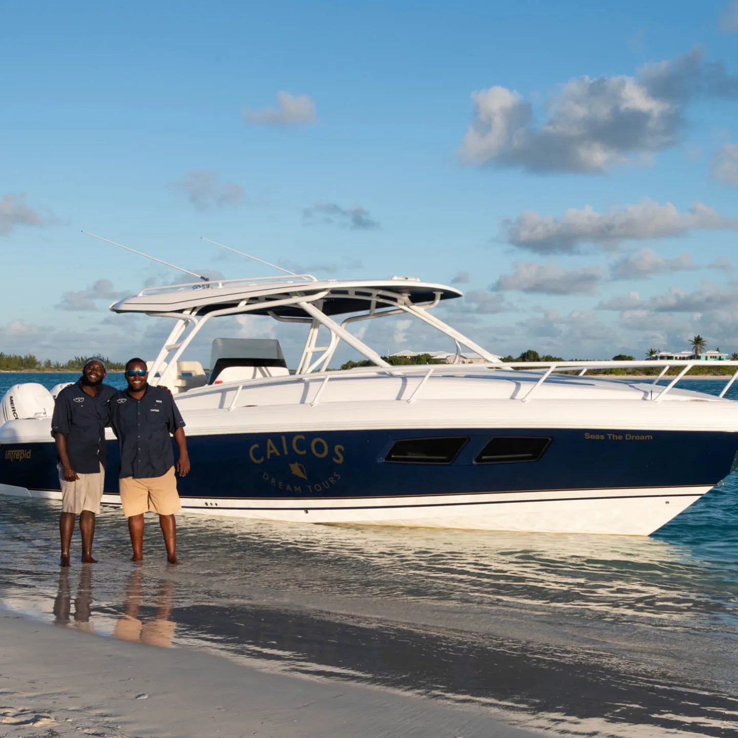 Two people stand on a beach near a white boat labeled 'Caicos' under a blue sky.