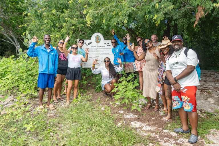 Group of people smiling and waving near a sign in a green, leafy area.