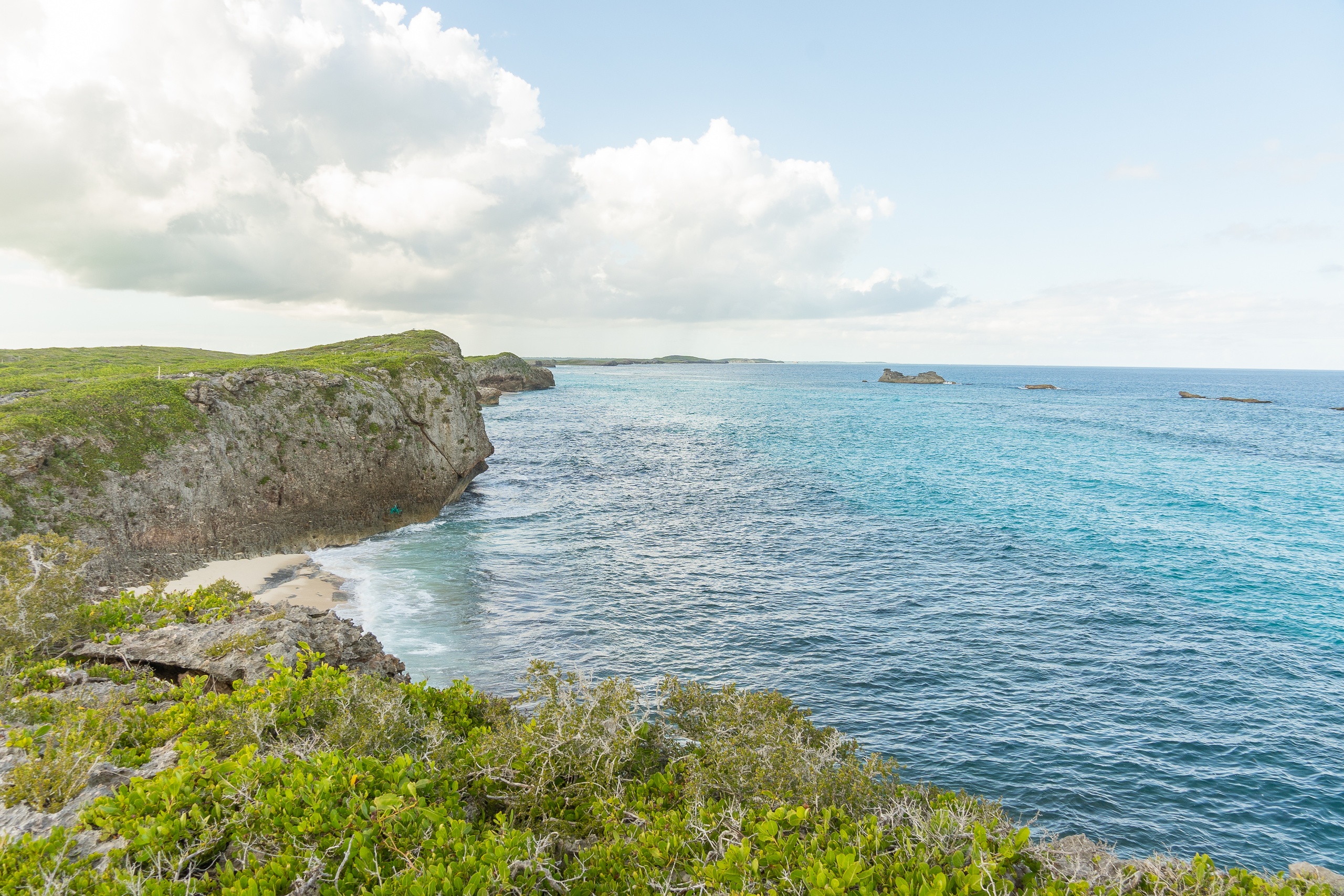 Coastal landscape with cliffs, green vegetation, and blue ocean under a partly cloudy sky.