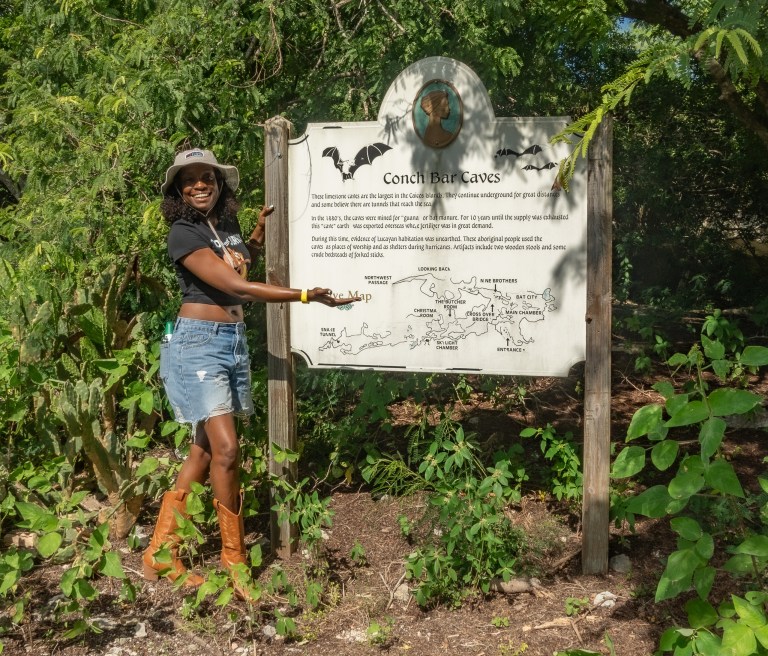 Woman in hat and boots points to sign about Conch Bar Caves in lush forest setting.