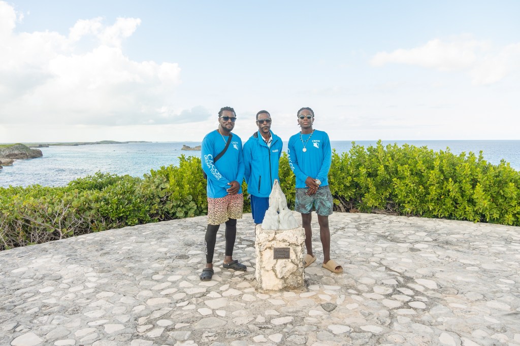 Three men in blue shirts stand on a stone patio by the ocean.