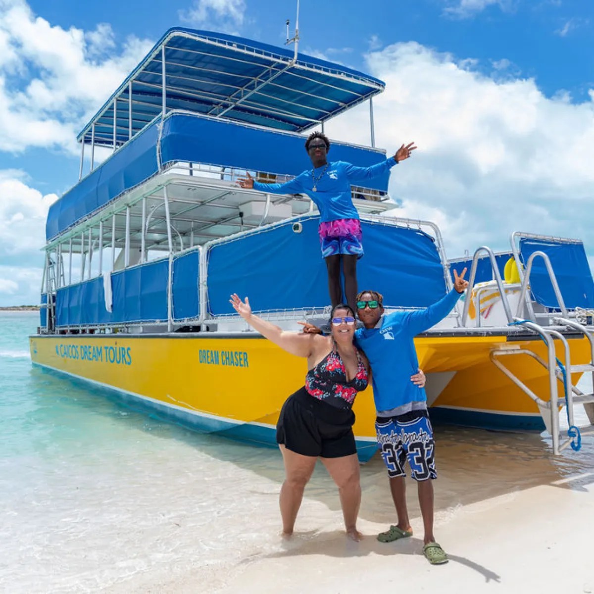 Three people posing joyfully by a blue and yellow tour boat on a beach.