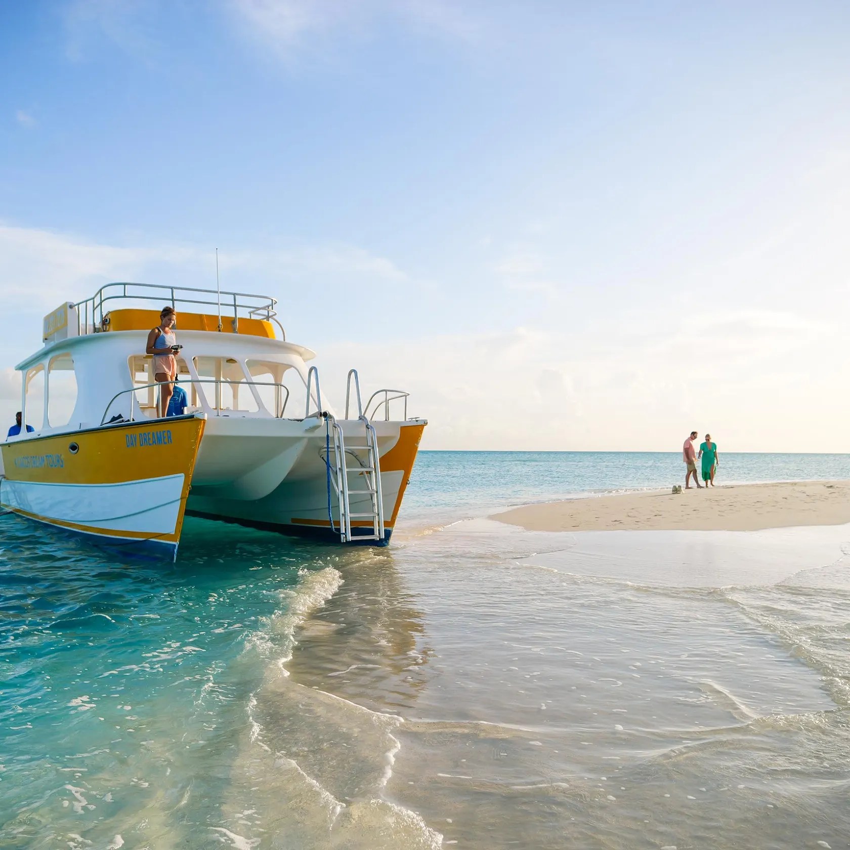Catamaran anchored near beach with people walking on sand.