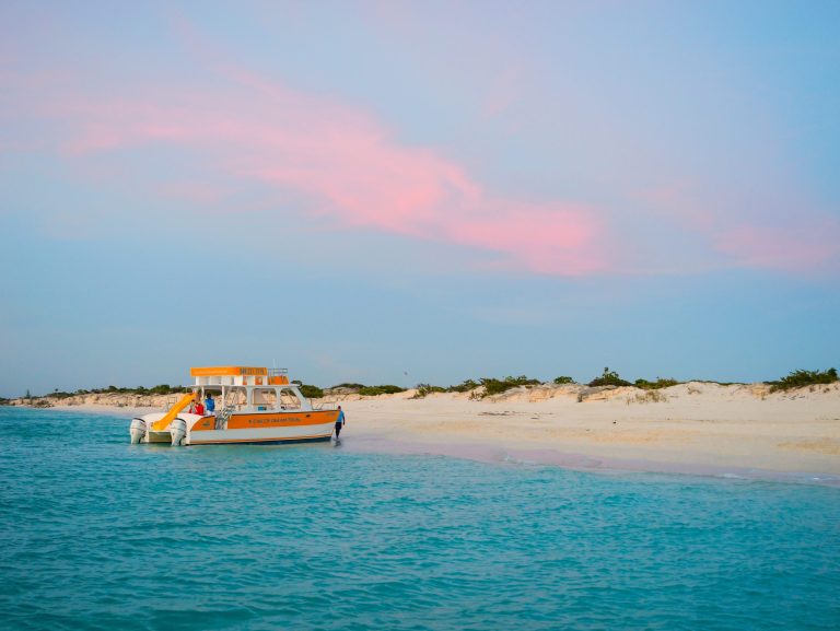 Boat on calm blue water near sandy beach under pink sky.