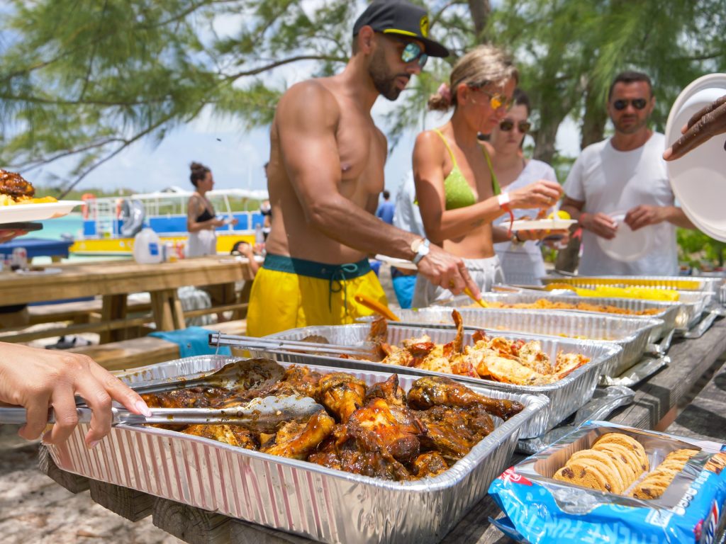 People serving food from trays at an outdoor buffet with trees and boats in the background.