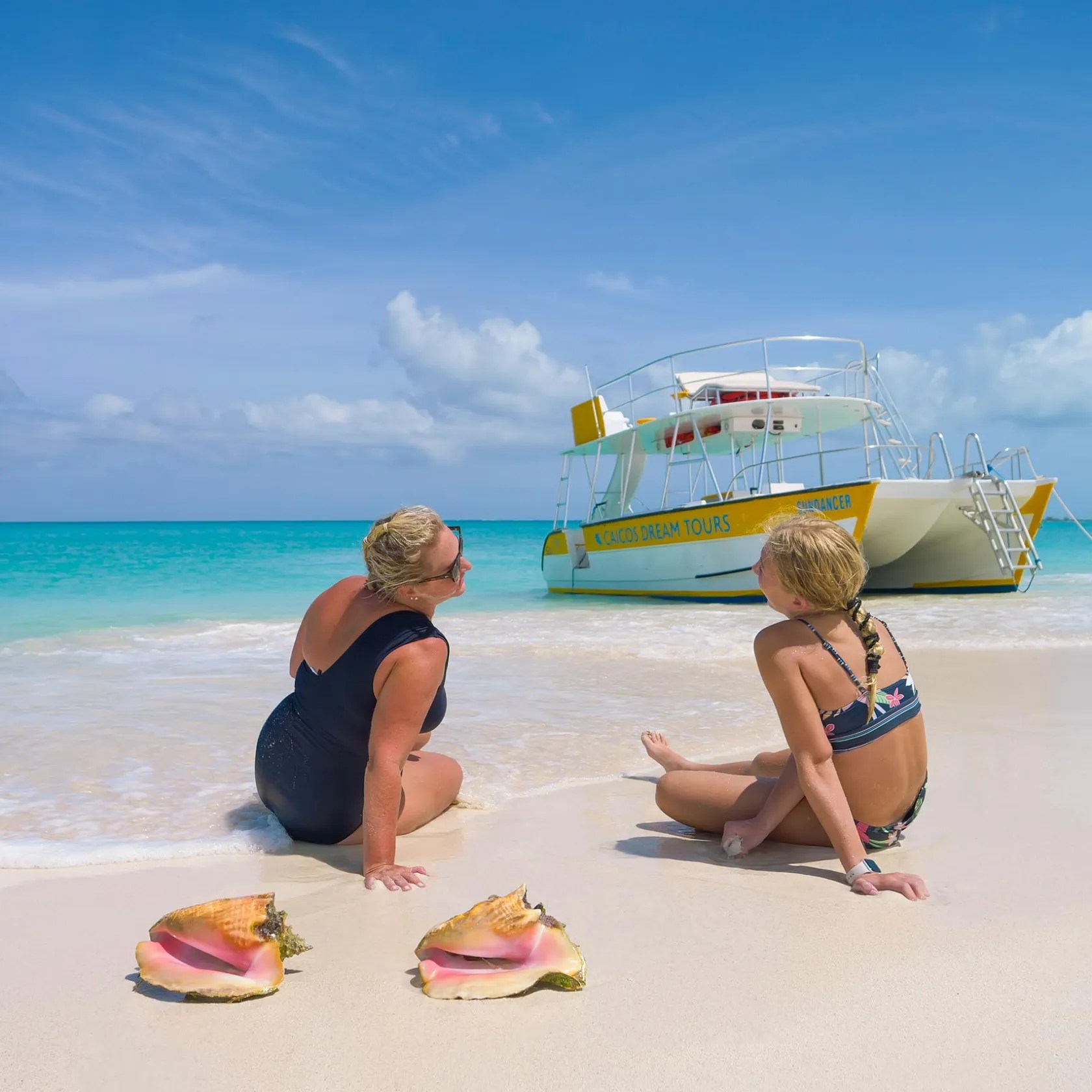 Two women sit on a sandy beach near large shells, with a boat on turquoise water in the background.
