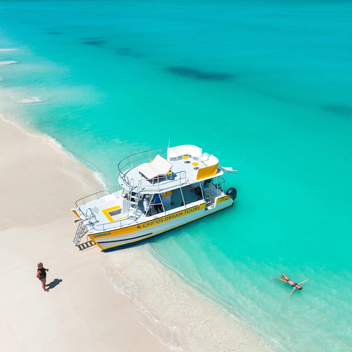 Boat on turquoise water near sandy beach, person swimming and another walking on shore.