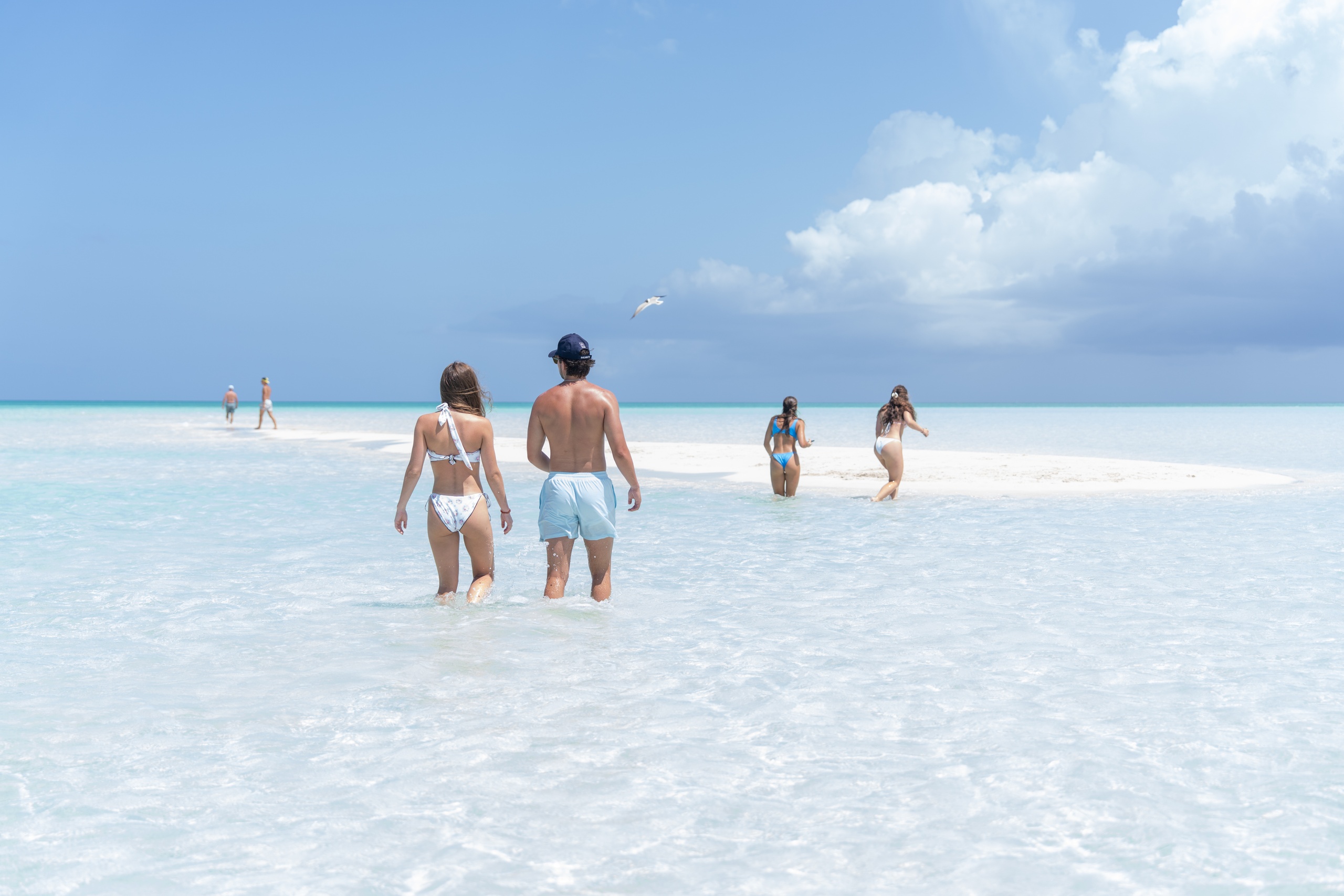 People walking in shallow, clear ocean water under a blue sky with scattered clouds.