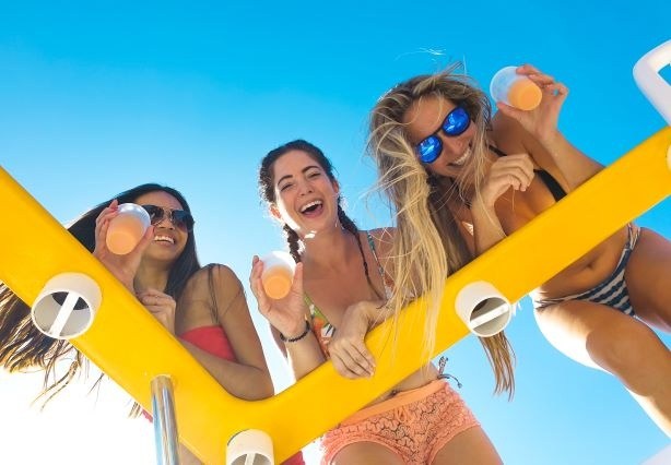 Three smiling women holding drinks above a yellow rail against a clear blue sky.
