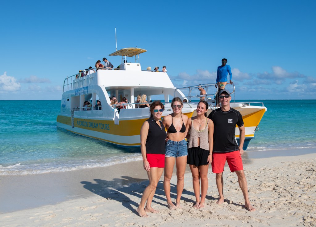 Four people posing on a sunny beach with a boat in the background.