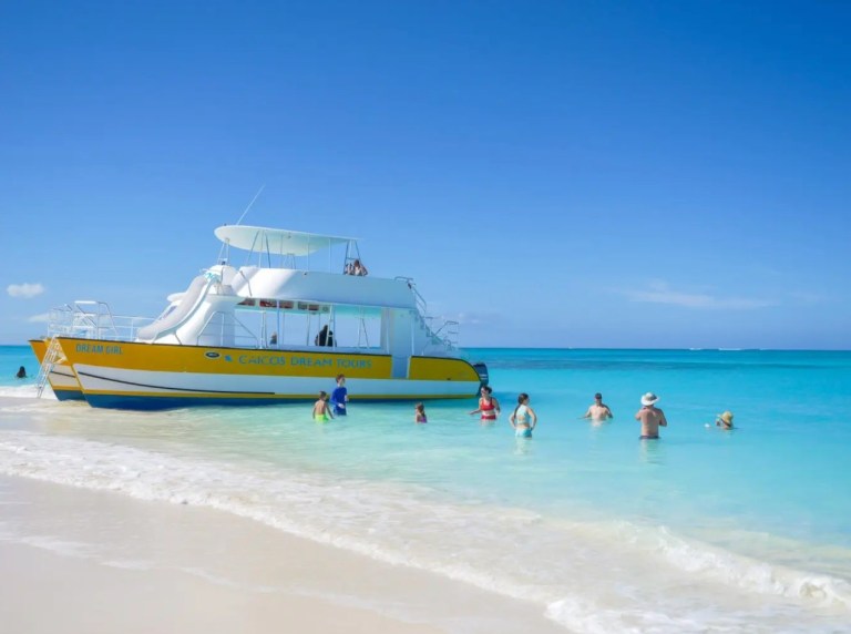 People wading in clear blue water near a white and yellow boat on a sunny day.