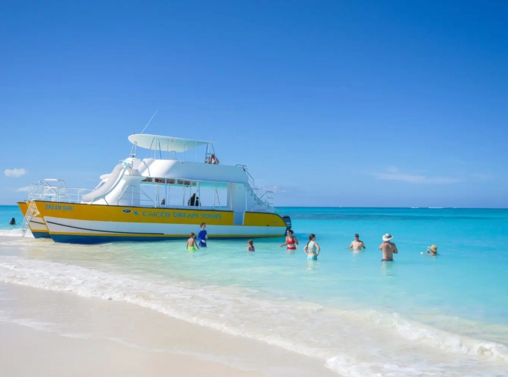 People wading in clear blue water near a white and yellow boat on a sunny day.