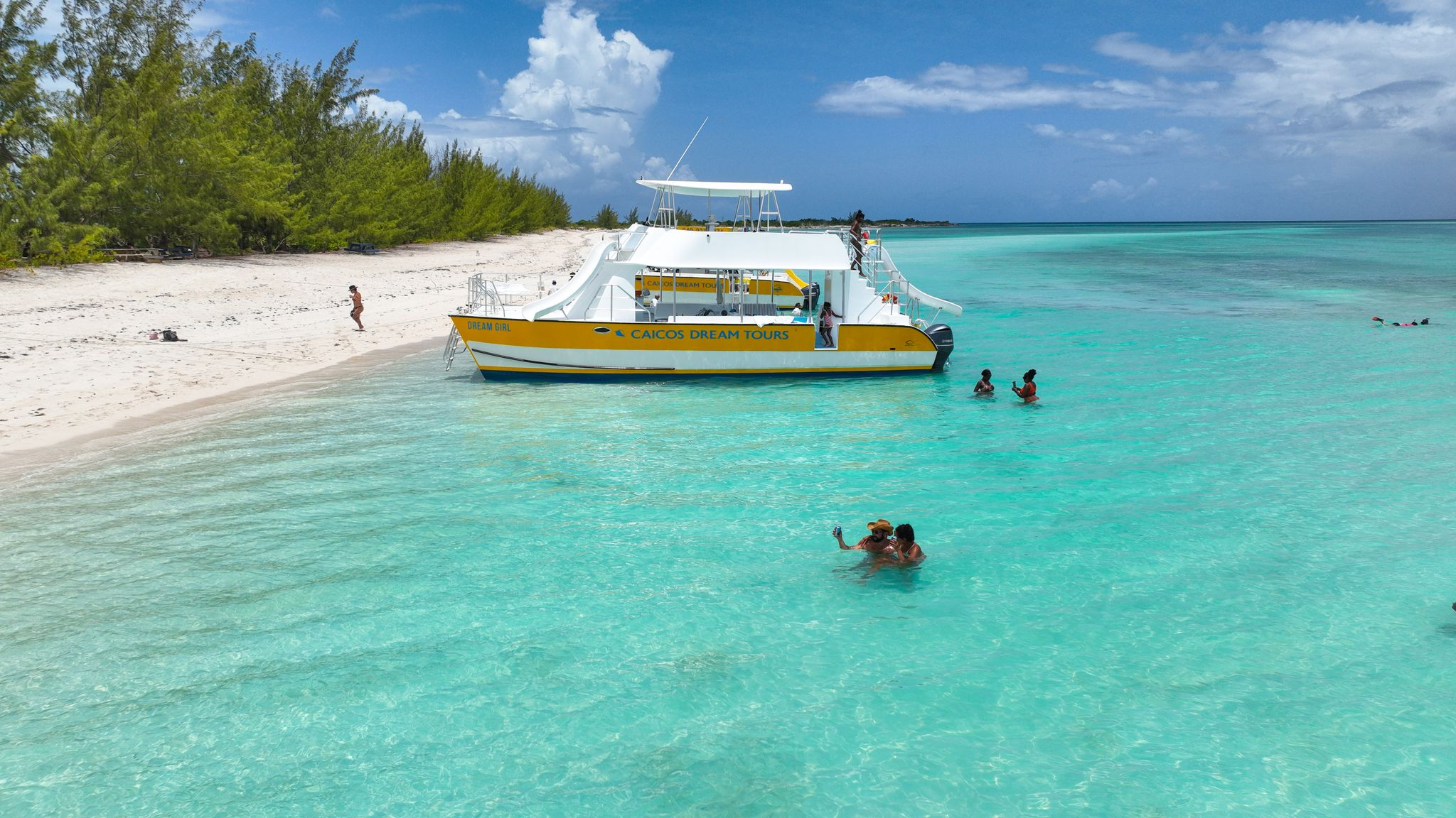 Yellow boat near a sandy beach with people swimming in turquoise water.