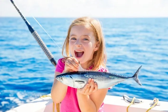 Smiling girl holding a fish on a boat with ocean in the background.
