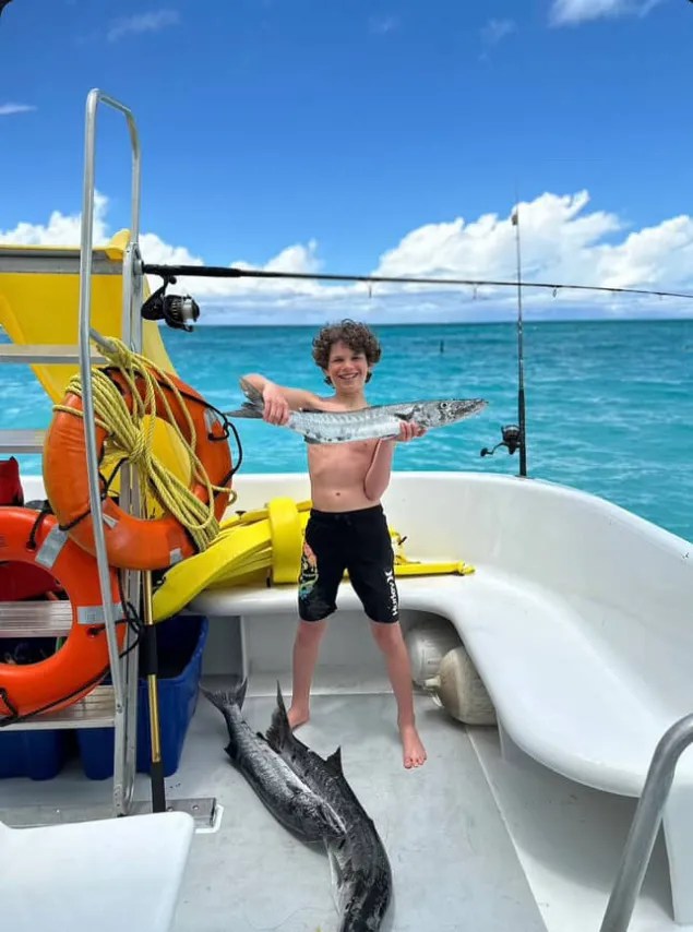 Boy on a boat holding a fish, two more fish on deck, blue ocean background.