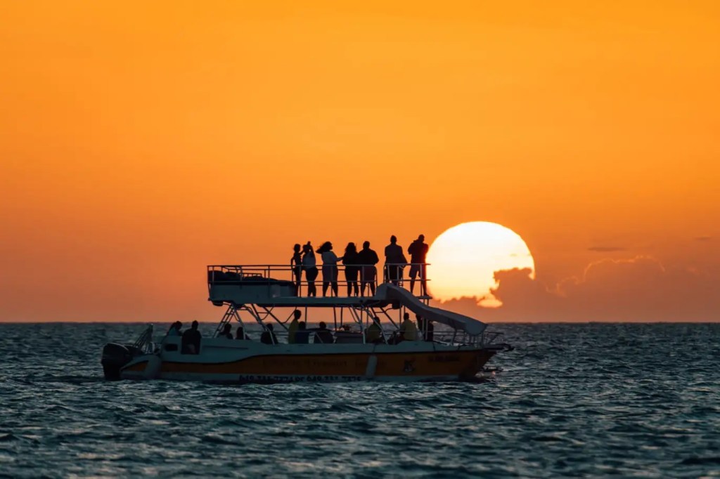 Silhouetted boat with people against a bright orange sunset over the ocean.