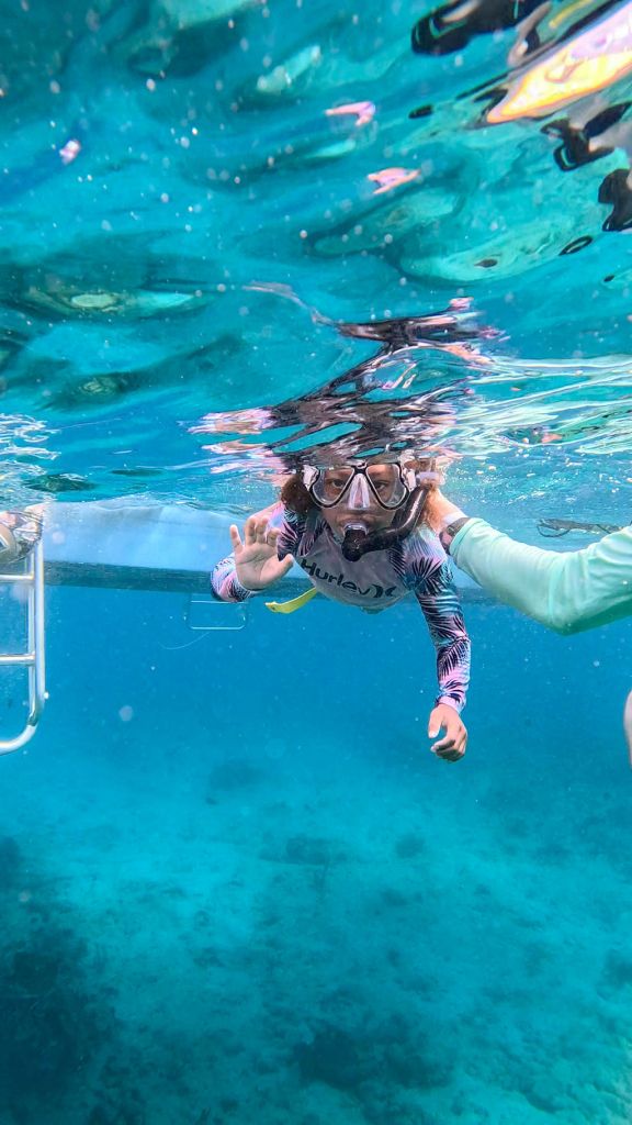 Person snorkeling underwater, reaching out, with clear blue water background.