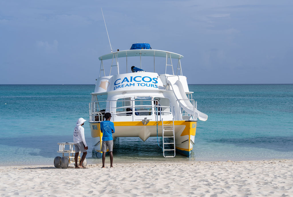 Boat on beach with 'Caicos Dream Tours' sign, people and ocean in background.