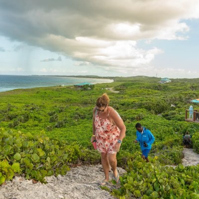 Woman in floral dress climbing a grassy hill with others, ocean and cloudy sky in background.
